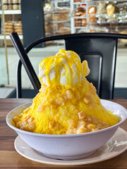 Ais Jagung, bright yellow shaved ice topped with vanilla ice cream, sweet corn and syrup, served in a bowl with fruit pieces and straws, placed on a wooden café table.