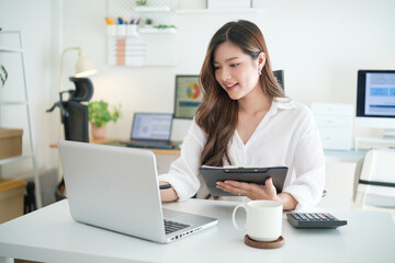 Smiling Asian woman working from home with a laptop and clipboard in a bright minimalist office.