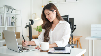 Young Asian woman writing notes while working on laptop at home office desk, smiling and focused.