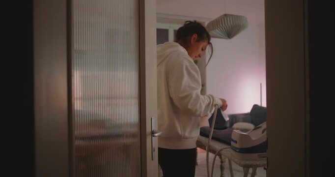 Woman ironing clothes in softly lit living room at night, surrounded by warmth of home and soft shadows, showing focus and care in everyday domestic life