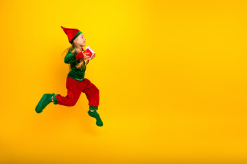 Cheerful elf kid jumping while carrying a gift against bright yellow background during festive Christmas season