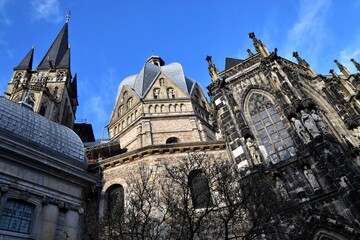 Low-angle view of the historic Aachen Cathedral in Germany, a UNESCO World Heritage site, showcasing its magnificent blend of Carolingian and Gothic architecture against a clear blue sky.

