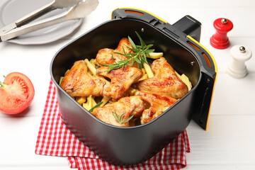 Grilled chicken wings with french fries in air fryer basket, tomato and tongs on white wooden table, closeup