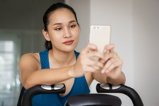 Distracted young real Asian woman uses smartphone while texting on a stationary exercise bike at a modern gym. This lifestyle image represents fitness, technology, multitasking, and healthy habits