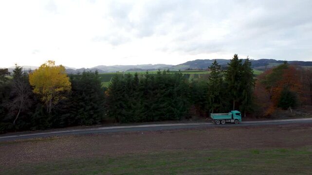 Baustellen-LKW auf Schotterstra&szlig;e entlang einer Baumreihe in herbstlicher Mittelgebirgslandschaft &ndash; Drohnenflug mit seitlicher Bewegung, Felder und untergehende Sonne im Hintergrund
