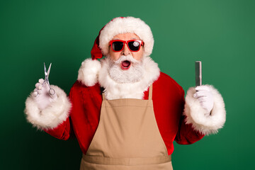 Cheerful Santa in red suit with apron holding scissors and comb in a festive green studio ready for holiday season and Christmas themed
