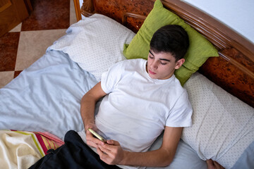 Teenage boy in white t-shirt sitting on bed, looking down at his smartphone while texting. Calm indoor moment in cozy bedroom.