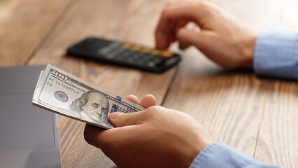 Man holding a pile of 100 dollar bills while using calculator. Financial planning, budgeting or tax calculation concept. Ideal for personal finance, business or investment content.