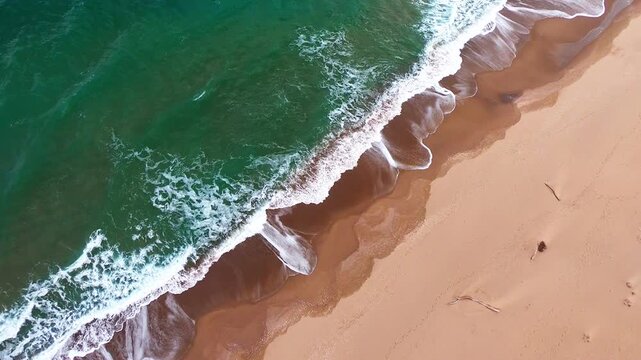 Drone video of a deserted wild beach and blue-green sea waves shot vertically from above, with the shoreline cutting diagonally across the frame
