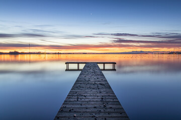 Naklejka premium Wooden pier in the Zuiderhaven of Den Oever during a colorful sunrise. The sun announces its arrival by painting the horizon orange, while the visible star remains as a reminder of the night.
