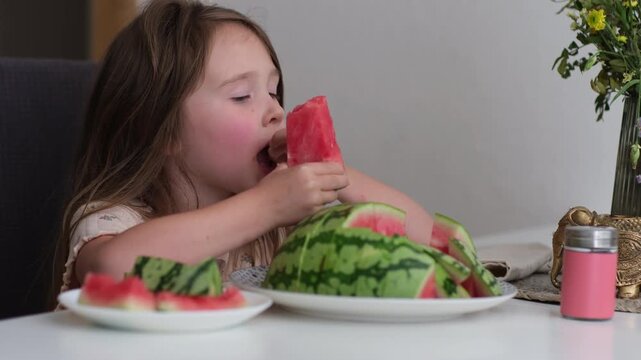 A small beautiful Caucasian girl sits at the table and eats a watermelon, making funny faces. There is a bouquet of flowers and a plate with crusts on the table. A cozy home atmosphere.