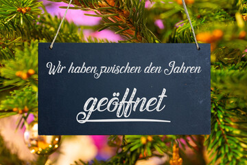 &Ouml;ffnungszeiten an Weihnachten: Schild mit der Aufschrift WIR HABEN ZWISCHEN DEN JAHREN GE&Ouml;FFNET vor einem Weihnachtsbaum