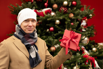 Happy man holding red gift box near Christmas tree. Xmas holiday spirit.