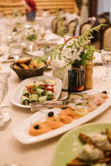 Elegant banquet table with appetizers: cucumber rolls, smoked salmon slices, tomato canapés, bread basket. Decorated with a floral arrangement in a dark bottle. Crystal glasses and tableware add