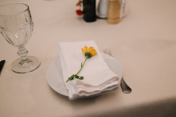 Elegant table setting with a white napkin folded on a plate, adorned with a delicate yellow flower. Accompanied by a crystal glass, fork, and subtle background condiments — perfect for a refined