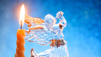 Glass angel figurine with trumpet and burning candle on shimmering blue background symbolizing...