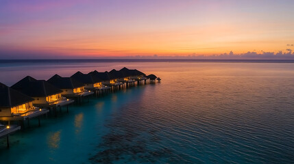 Obraz premium A tropical overwater bungalows stand gracefully above the turquoise sea at sunset, casting a warm glow as the sky blends into a stunning array of orange and purple hues.