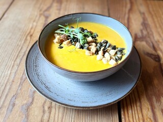 Creamy pumpkin soup served in a grey bowl, topped with croutons, toasted pumpkin seeds and fresh herbs, placed on a wooden table in warm natural light.