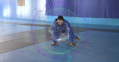 Performing martial artist wearing blue gi crouching low on mat in gym, with digital hologram sphere