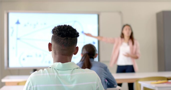 Watching teenage boy and girl sitting at desks in classroom, teacher pointing at graph display