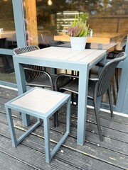 Outdoor café table with modern chairs and a small matching stool, featuring a potted plant centerpiece on a wooden deck beside a glass window.