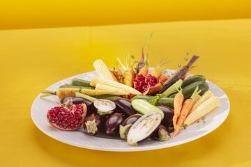 A plate of assorted raw vegetables like eggplant, carrots, cucumber, and baby corn is placed with pomegranate sections on a white plate against a solid yellow background