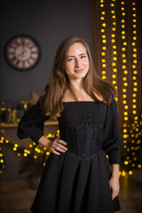 A woman in a black dress and stockings stands in front of a Christmas tree. She smiles and poses for the camera. The concept of holidays and Christmas.