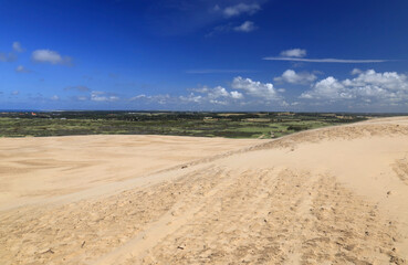 Landscape photo with a view of sand dunes in the foreground against a blue sky with clouds near the Rubjerg Knude lighthouse in Denmark