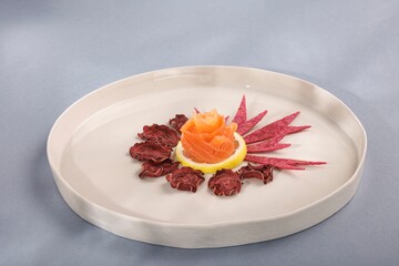Delicate salmon crafted into a rose sits upon a lemon slice. Around it, purple chips arranged like petals and leaves, displayed on a stark white plate