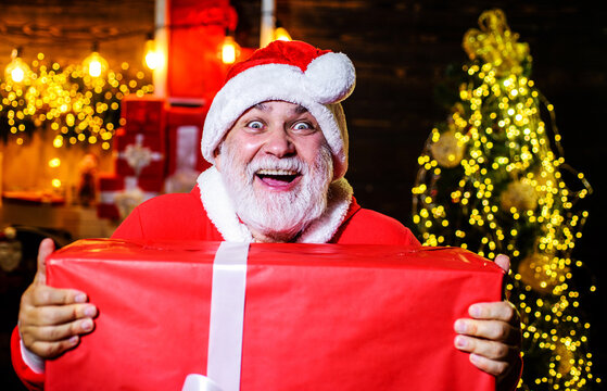 Smiling Santa Claus with large Christmas gift box. Bearded man in Santa Claus wear with big Christmas present. Festive New Year decorations. Christmas and New Year sales. Delivery service. Gift shop.