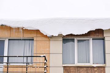 Melting snow overhang on residential roof, icicle hazard during thaw and winter safety risk in urban area. Building facade with heavy roof snow accumulation, frozen snowpack on roof edge above window