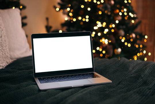 Open laptop with blank screen on cozy bed, Christmas tree with warm lights in the background, festive home atmosphere. - Powered by Adobe