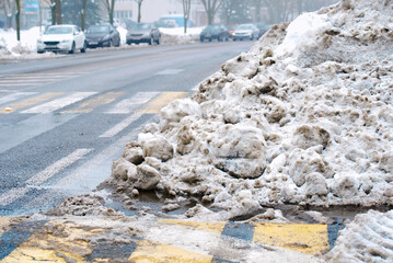 Frozen melting snow near pedestrian area, municipal cleanup aftermath. Dirty slushy pile on asphalt surface, melting ice and thawing runoff near street crossing. Street cleaning remains, melting snow