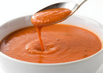 Smooth tomato soup dripping from a silver spoon into a white bowl on a clean surface