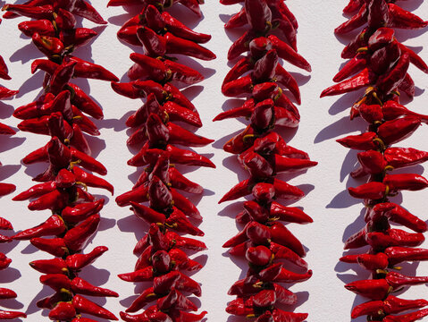 Rows of bright red Espelette peppers dry in midday sun against a white wall, casting graphic shadows in Espelette, France.