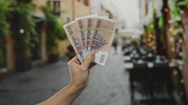 Man holding icelandic krona banknotes on a city street terrace with blurred outdoor restaurant setting, capturing economic and travel themes.