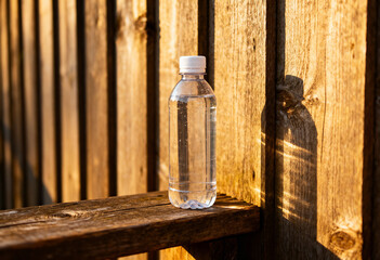 A clear plastic water bottle with a white cap sits on a wooden surface outdoors in sunlight