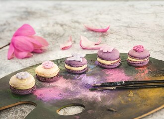 A selection of pastel macarons, each decorated with tiny flowers, rests on a paint palette with brushes. Petals from a pink flower add a touch of springtime beauty to this sweet scene