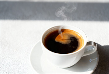 A close up of a white coffee cup filled with dark coffee and rising steam on a white surface