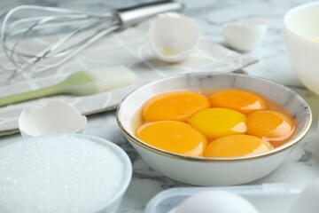 Broken eggs, yolks, sugar, brush and whisk on white marble table, closeup