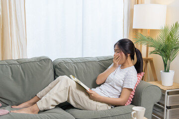 Fototapeta premium Young woman teen person reading book on sofa finds rest in comfortable living room, yawning subtly enjoying peaceful leisure time at home, cozy comfortable atmosphere.