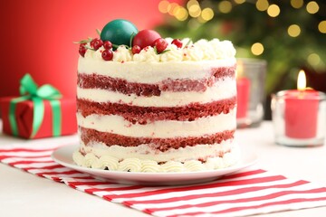Delicious sponge cake with Christmas decor on white table against red background with blurred lights, closeup. Bokeh effect