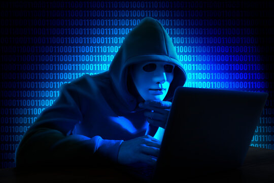 Cyber attack. Hacker in mask using laptop. Blue glowing binary code behind him