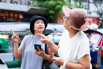 Senior asian women tourists looking up and pointing while checking smartphone near tuk tuk during travel city vacation symbolizing friendship exploration adventure and retirement lifestyle