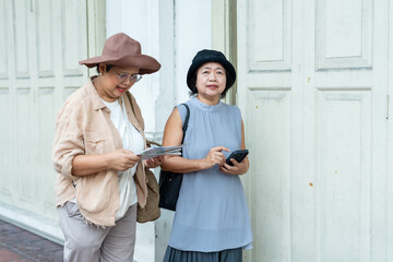Senior asian female friends travelers standing near white wall holding map and smartphone discussing travel plan enjoying retirement lifestyle vacation urban exploration with happiness and friendship