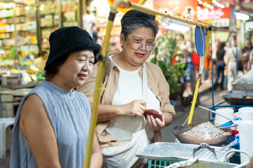 Senior asian female travelers buying street dessert smiling enjoying local food culture vacation lifestyle friendship during retirement exploring city market urban travel evening journey happiness