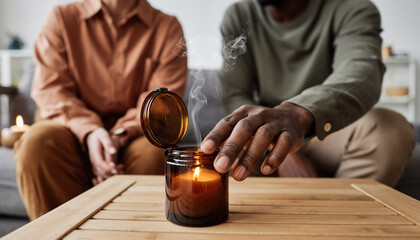 Diverse couple creating a cozy atmosphere at home, with a man's hand opening a warm, smoking aromatherapy candle for relaxation