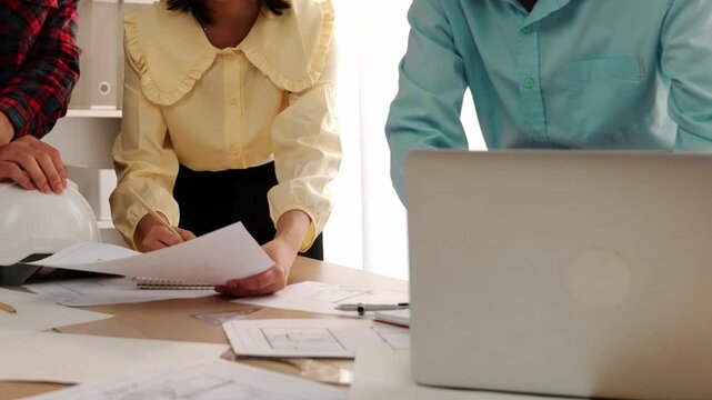 Architect and safety engineer discussion about building blueprint displaying on laptop on table with architectural document and wooden block scatter around at modern office. Closeup. Delineation.