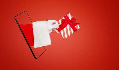 Christmas Santa Claus hand holding or gives red gift box with red bow from smartphone or mobile phone on red background with copy space. Design element. Banner.