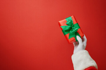 Christmas Santa Claus hand holding red striped gift box with green ribbon on festive red background with copy space for text, Christmas card design element.